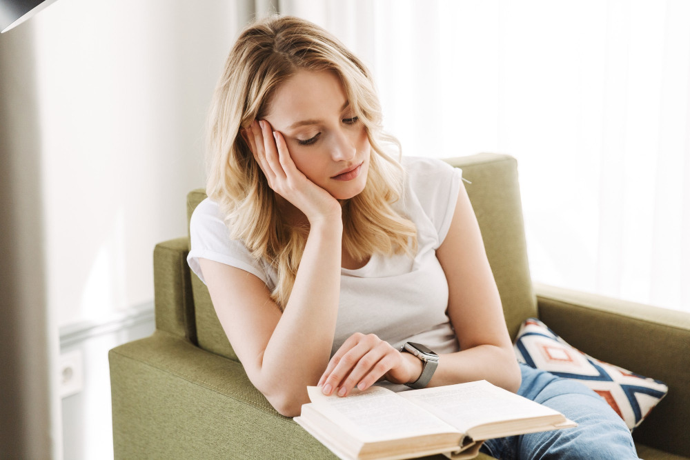 Junge Frau sitzt nachdenklich in einem grünen Sessel und liest ein offenes Buch in einem hellen Wohnraum.