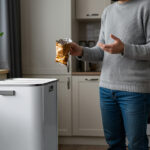 Eine Person steht zu Hause vor einer Recycling-Tonne und hält eine leere Snack-Verpackung ohne Recyclinghinweise in der Hand.