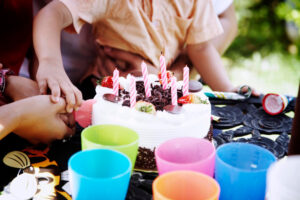 Kindergeburtstag mit bunter Torte und Kerzen, umgeben von farbigen Bechern, Kinderhände greifen nach dem Kuchen – fröhliche Feieratmosphäre.