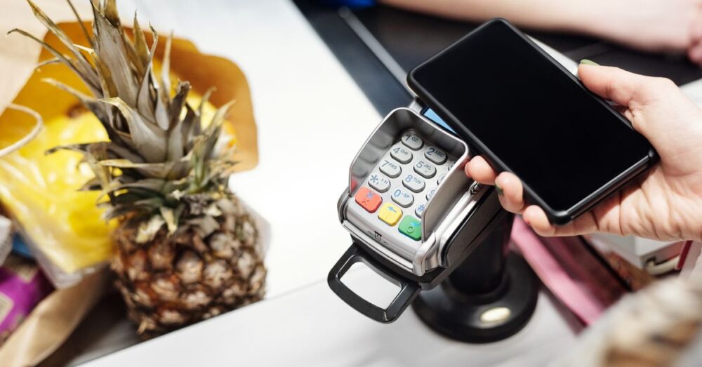 Hand hält ein Smartphone zum kontaktlosen Bezahlen an ein Kartenterminal; daneben liegt eine Ananas auf dem Kassentresen.