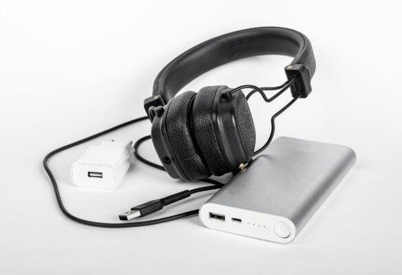 Schwarze Bügelkopfhörer liegen auf einer silbernen Powerbank; daneben liegen ein weißes Netzteil und schwarze Kabel.