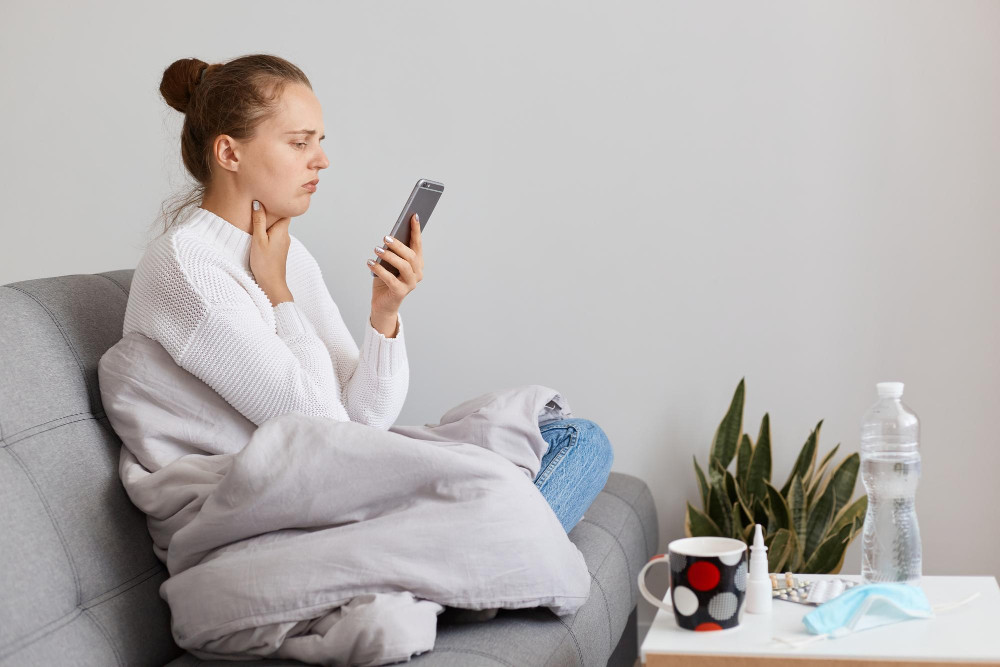 Eine sichtlich erkältete Frau sitzt mit Halsschmerzen und in eine Decke gewickelt auf dem Sofa und sucht besorgt auf ihrem Smartphone nach ärztlichem Rat oder der Nummer des Bereitschaftsdienstes; auf dem Beistelltisch stehen Wasser, Nasenspray und Medikamente bereit.