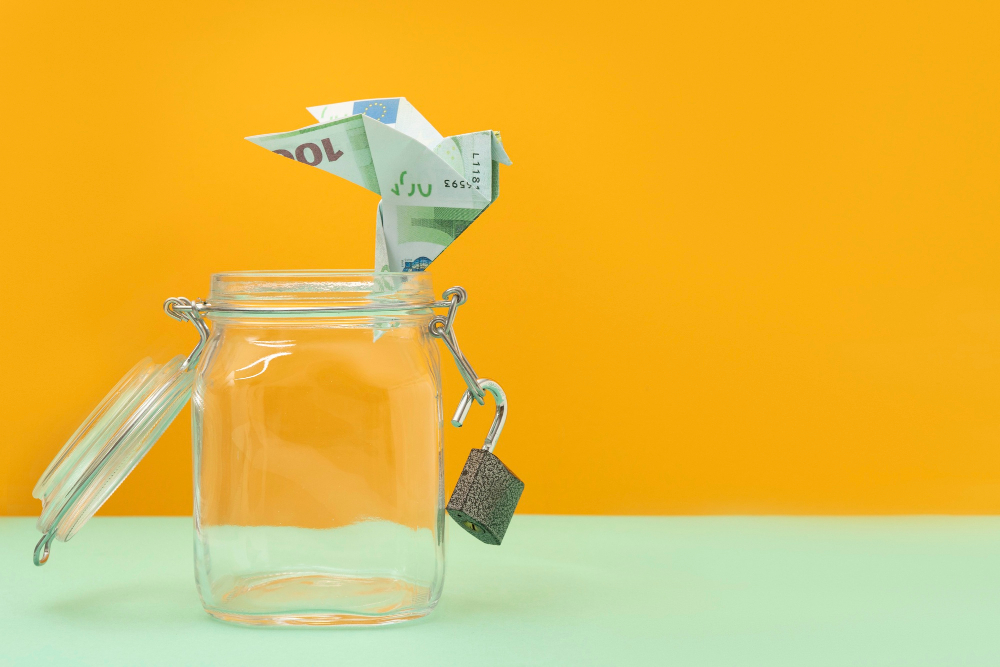 Ein zu einem Vogel gefalteter 100-Euro-Schein sitzt auf dem Rand eines leeren, offenen Einmachglases, das als Spardose dient. Das Bild vor einem leuchtend orangen Hintergrund symbolisiert kreative und ungewöhnliche Wege, Geld zu sparen.