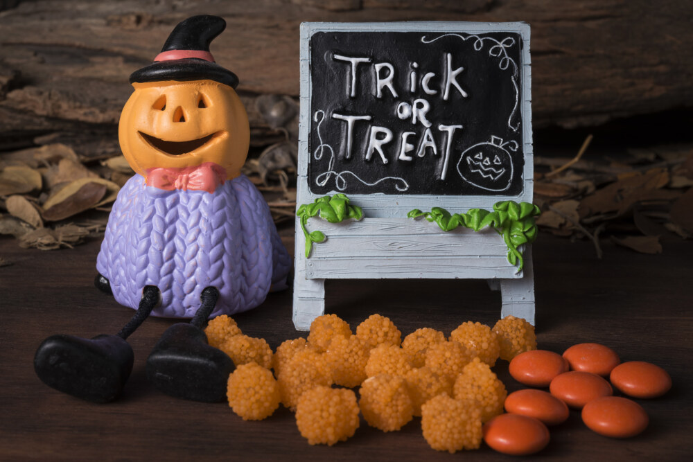 Eine Kürbisfigur mit schwarzem Hut sitzt neben einer Tafel mit „Trick or Treat“; davor liegen orange Süßigkeiten.