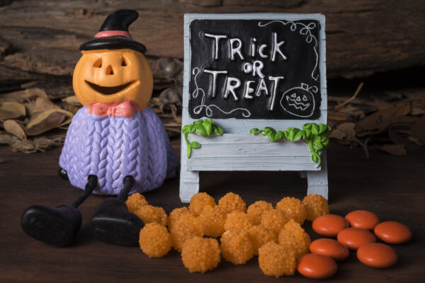 Eine Kürbisfigur mit schwarzem Hut sitzt neben einer Tafel mit „Trick or Treat“; davor liegen orange Süßigkeiten.