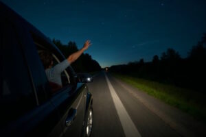 Ein Auto fährt nachts über eine ruhige Landstraße in Deutschland. Eine Person streckt den Arm aus dem heruntergelassenen Fenster in Richtung des dunklen Himmels, an dem vereinzelt Sterne zu sehen sind. Die Szene vermittelt ein Gefühl von Freiheit, Sommerstimmung und entspanntem Late-Night-Drive.