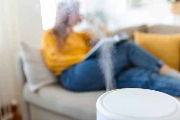 Ein weißes Gerät gibt feinen Nebel ab, während eine Frau in gelbem Oberteil und Jeans auf dem Sofa sitzt.