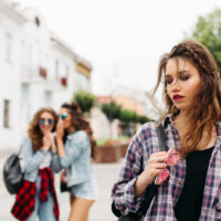Junge Frau mit pinker Sonnenbrille schaut nachdenklich zur Seite, zwei andere Mädchen im Hintergrund flüstern