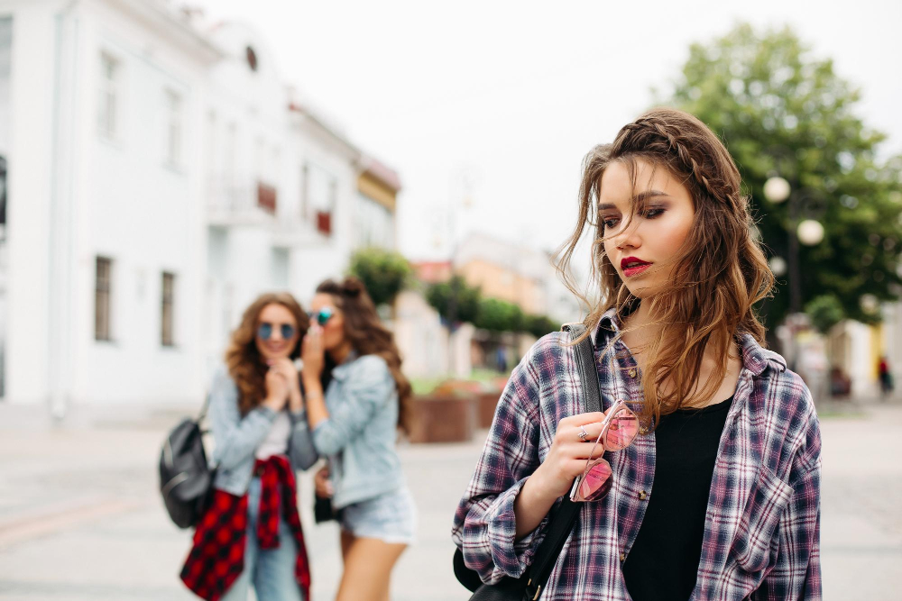 Junge Frau mit pinker Sonnenbrille schaut nachdenklich zur Seite, zwei andere Mädchen im Hintergrund flüstern