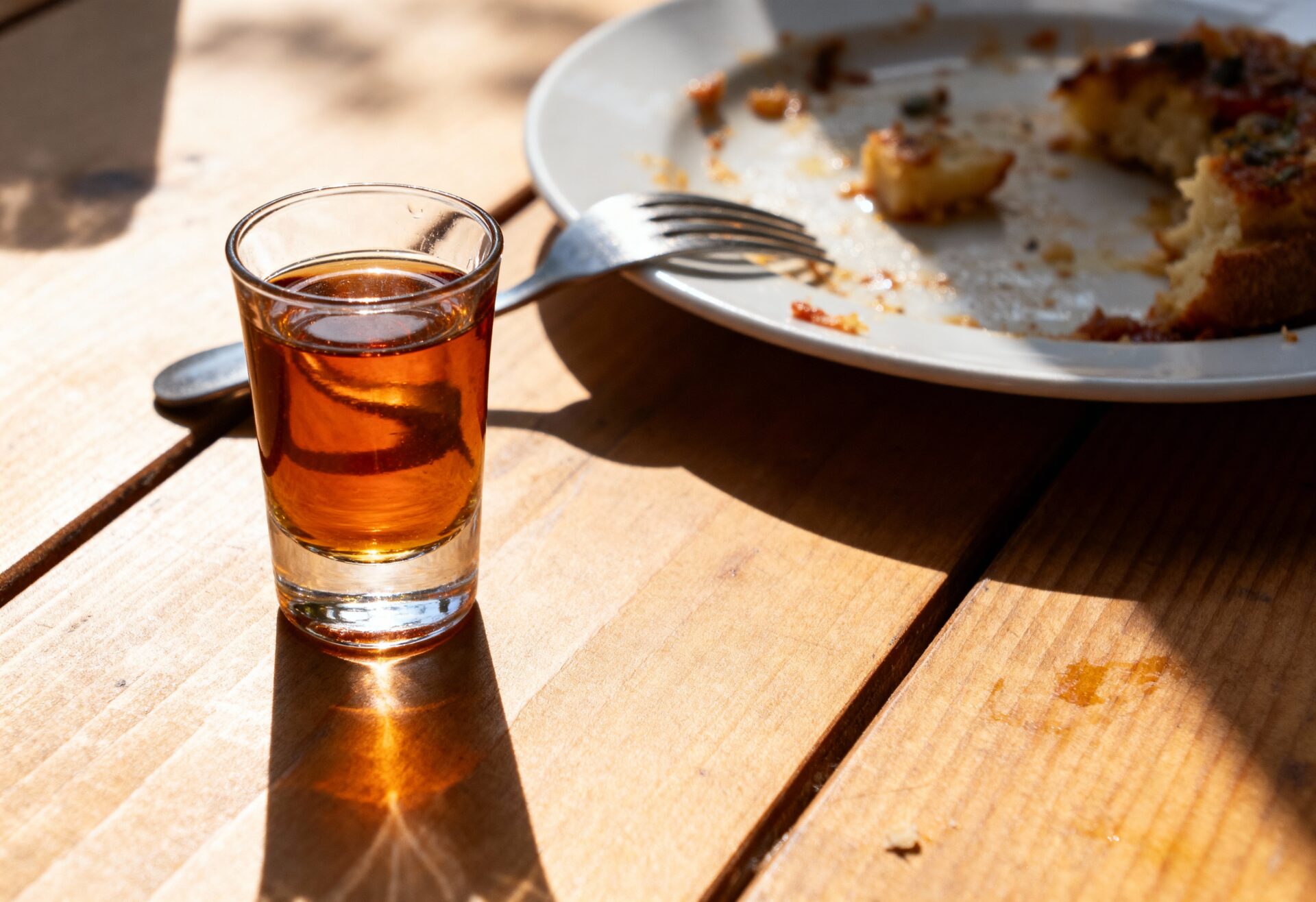 Kleines Glas mit bernsteinfarbenem Kräuterbitter auf Holztisch, daneben Teller mit Resten vom Essen und Gabel im Sonnenlicht.