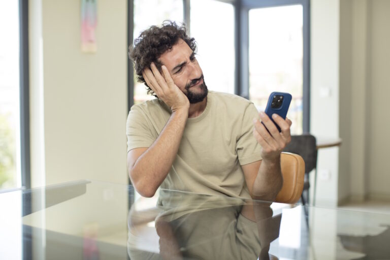 Mann mit lockigem Haar stützt den Kopf und blickt genervt auf sein Smartphone