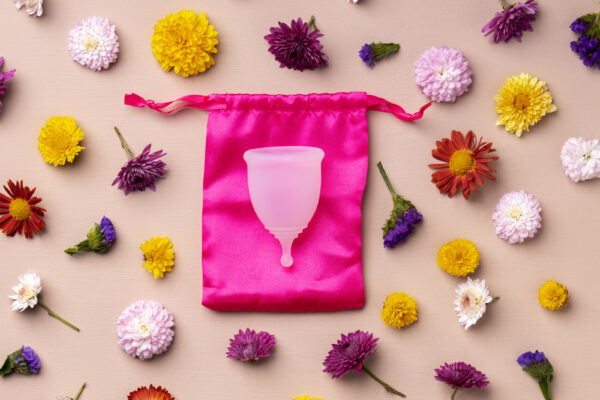 Eine blassrosa Menstruationstasse liegt auf einem kräftig pinken Stoffbeutel, der von bunten Blumen umgeben ist.