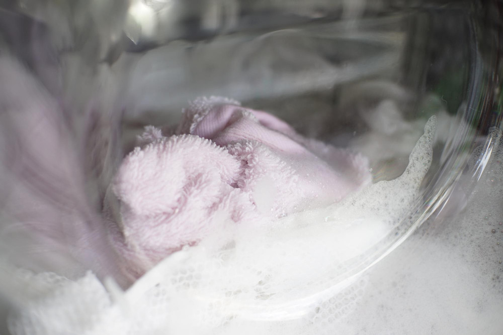 Mikrofaser richtig waschen: rosa Mikrofasertuch im Waschnetz in der Waschmaschine, sanfter Schaum beim Schonwaschgang.