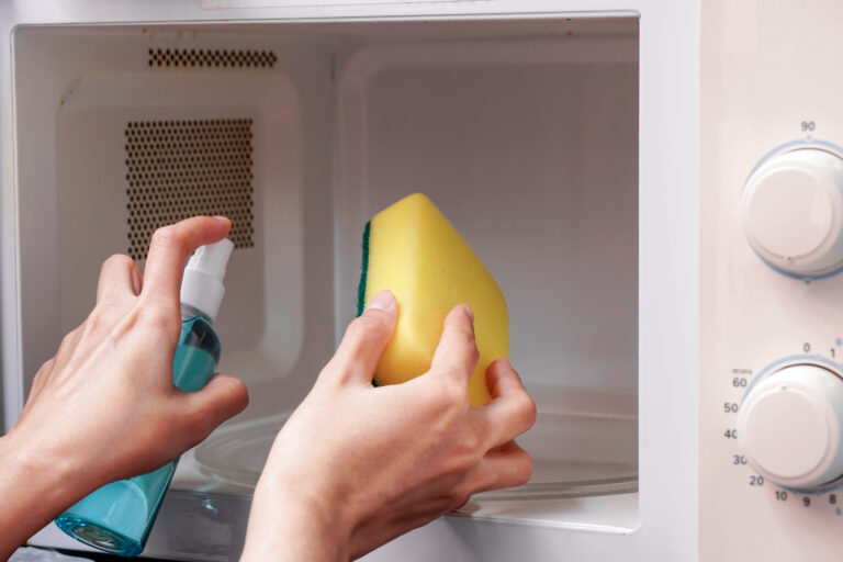 Hand reinigt den Innenraum einer Mikrowelle mit Schwamm und Reinigungsspray