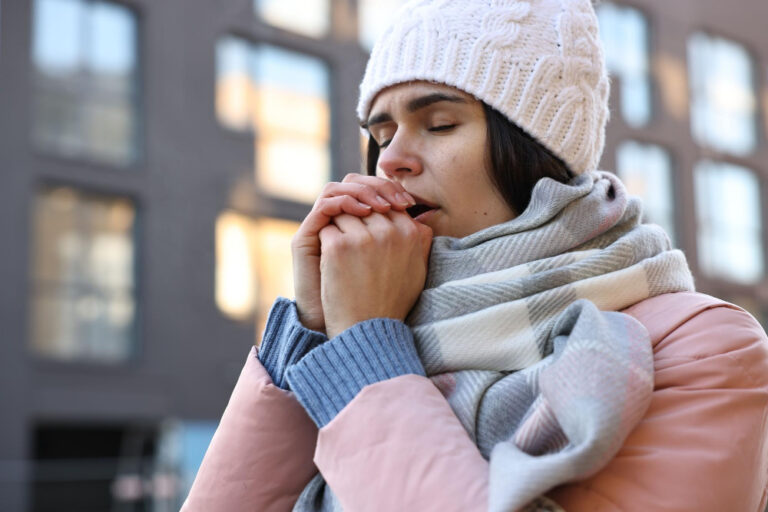Eine Frau in Winterkleidung mit Mütze und Schal steht draußen und hält ihre Hände an den Mund, als ob sie versucht, sich zu wärmen. Das Bild symbolisiert das Thema 'Mir ist die ganze Zeit kalt' und zeigt das Gefühl von Kälte trotz warmer Kleidung.