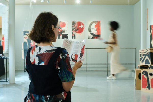 Eine Frau steht von hinten in einer Kunstgalerie und liest konzentriert einen Ausstellungs-Flyer, während im Hintergrund eine andere Besucherin an Gemälden vorbeigeht.