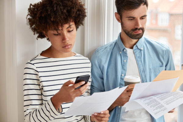 Online-Banking Fehlüberweisung zurückholen: junges Paar prüft Rechnungen am Fenster, Frau vergleicht Daten am Smartphone - symbolisiert schnelle Reaktion und Kontrolle.