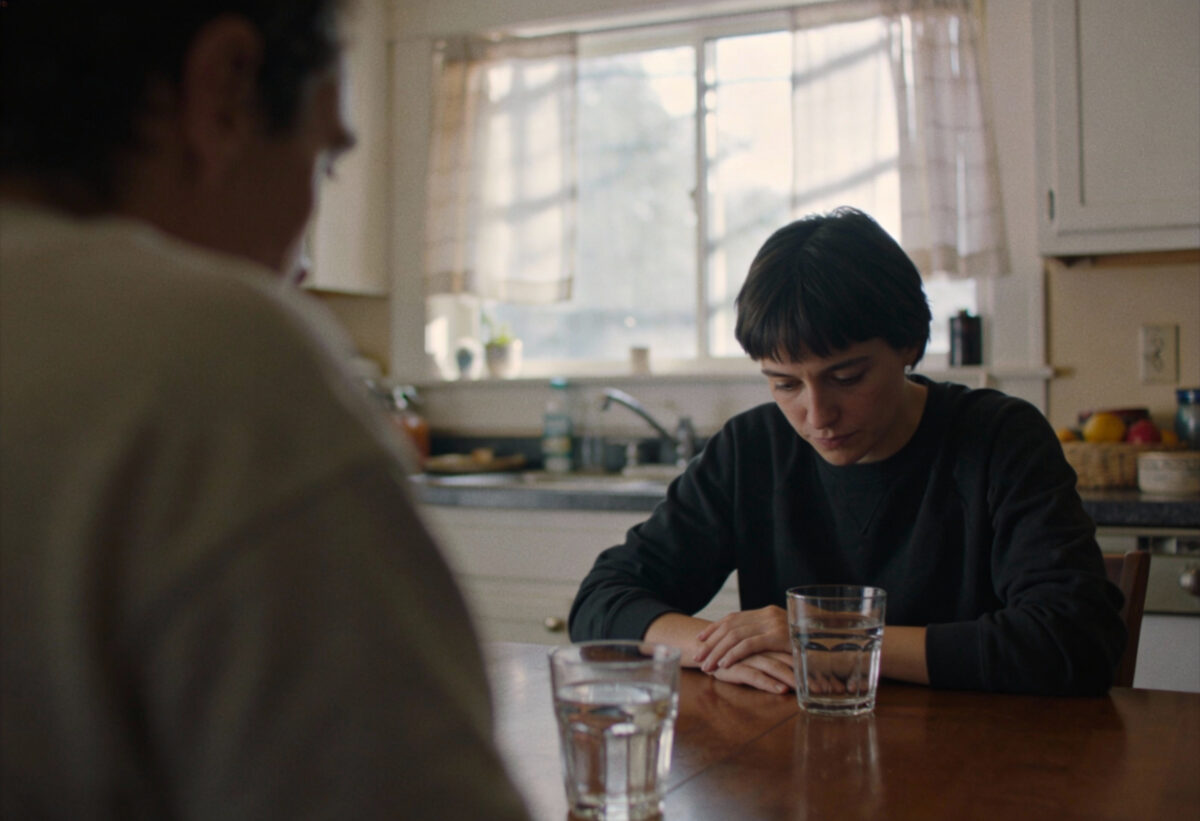 Zwei Personen am Küchentisch in peinlicher Gesprächspause, beide schauen verlegen auf ihre Wassergläser