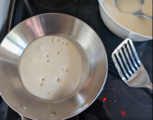 Erster Pfannkuchen: dünne Teigschicht in heißer Edelstahlpfanne, etwas Öl verteilt; daneben Schüssel mit Teig und Metallwender.