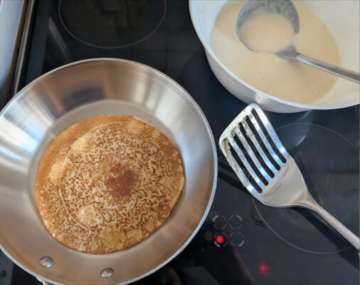 Goldbraun gebackener Pfannkuchen nach dem Wenden in heißer Edelstahlpfanne; nichts klebt, daneben Teigschüssel und Metallwender.