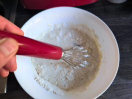 Hand mit rotem Schneebesen rührt Mehl-Milch-Ei in einer weißen Schüssel zu Pfannkuchenteig an.