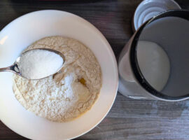 Löffel mit Zucker über einer Schüssel mit Mehl, Ei und Milch - Zutaten für Pfannkuchenteig vor dem Verrühren.