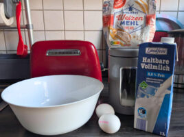 Pfannkuchen-Zutaten in der Küche: weiße Rührschüssel, zwei Eier, Milchpackung, Mehl, Zuckerbehälter und Handmixer - bereit zum Anrühren des Grundteigs.