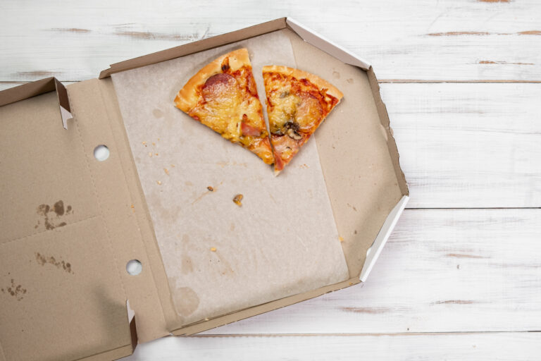 In einem offenen braunen Pizzakarton liegen zwei Pizzastücke; daneben sind Krümel und Fettspuren zu sehen.