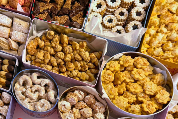 Mehrere Keksdosen mit unterschiedlichen Weihnachtsplätzchen, darunter Vanillekipferl, Schoko-Ecken, Spitzbuben und Spritzgebäck, dicht gefüllt auf einem Tisch.