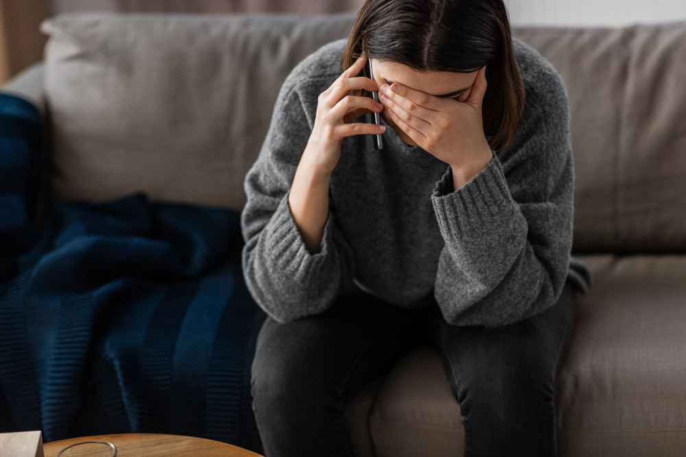 Eine Frau sitzt auf einem grauen Sofa, hält sich die Hand vor das Gesicht und telefoniert – Darstellung eines emotionalen Moments mit plötzlichem Weinen.