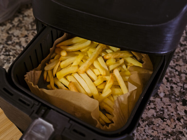Ein geöffneter schwarzer Airfryer mit gelb-goldenen Pommes auf braunem Backpapier im Korb.