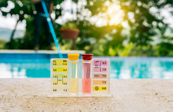Ein Pool-Wassertester mit zwei Röhrchen steht am Beckenrand; dahinter ist blaues Wasser zu sehen.