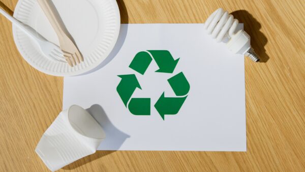 Grünes Recyclingzeichen auf einem weißen Blatt, daneben Einweggeschirr, Besteck, ein Becher und eine Energiesparlampe.