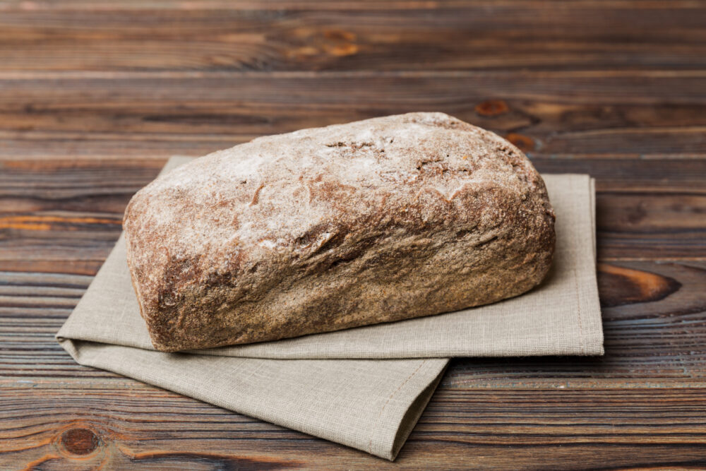 Ein rustikaler, hell bemehlter Brotlaib liegt auf einem gefalteten beigefarbenen Tuch auf einem dunklen Holztisch.