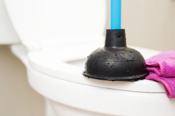 Eine schwarze Saugglocke mit blauem Griff liegt auf dem weißen Toilettensitz, daneben steckt ein pinkes Tuch.