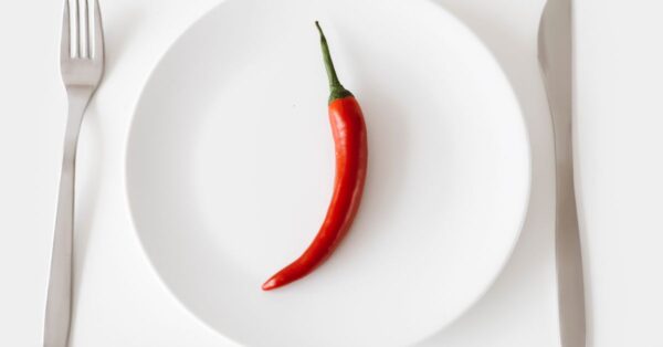 A single red chili pepper on a white plate with fork and knife, embodying simplicity and spicy flavor.
