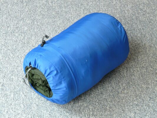 Blauer Schlafsack zusammengerollt in seinem Packsack auf grauem Untergrund