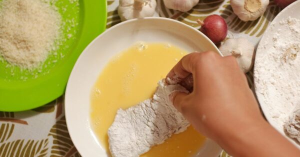 Hand zieht ein paniertes Schnitzel durch verquirltes Ei, daneben stehen Mehl und Semmelbrösel