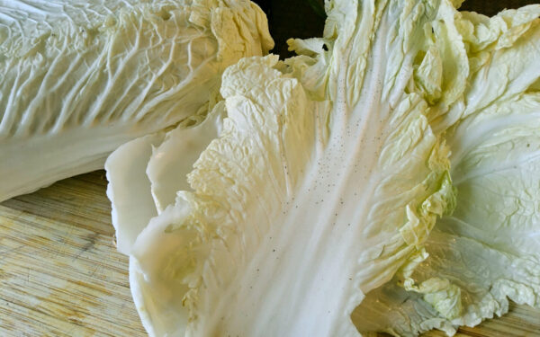 Nahaufnahme eines Chinakohlblatts, das deutliche Anzeichen von kleinen, dunklen "Pfeffenflecken" (physiologische Störung) auf der weißen Mittelrippe und den hellgrünen Bereichen aufweist. Im Hintergrund sind weitere Blätter des Chinakohls auf einem Holzschneidebrett zu sehen.