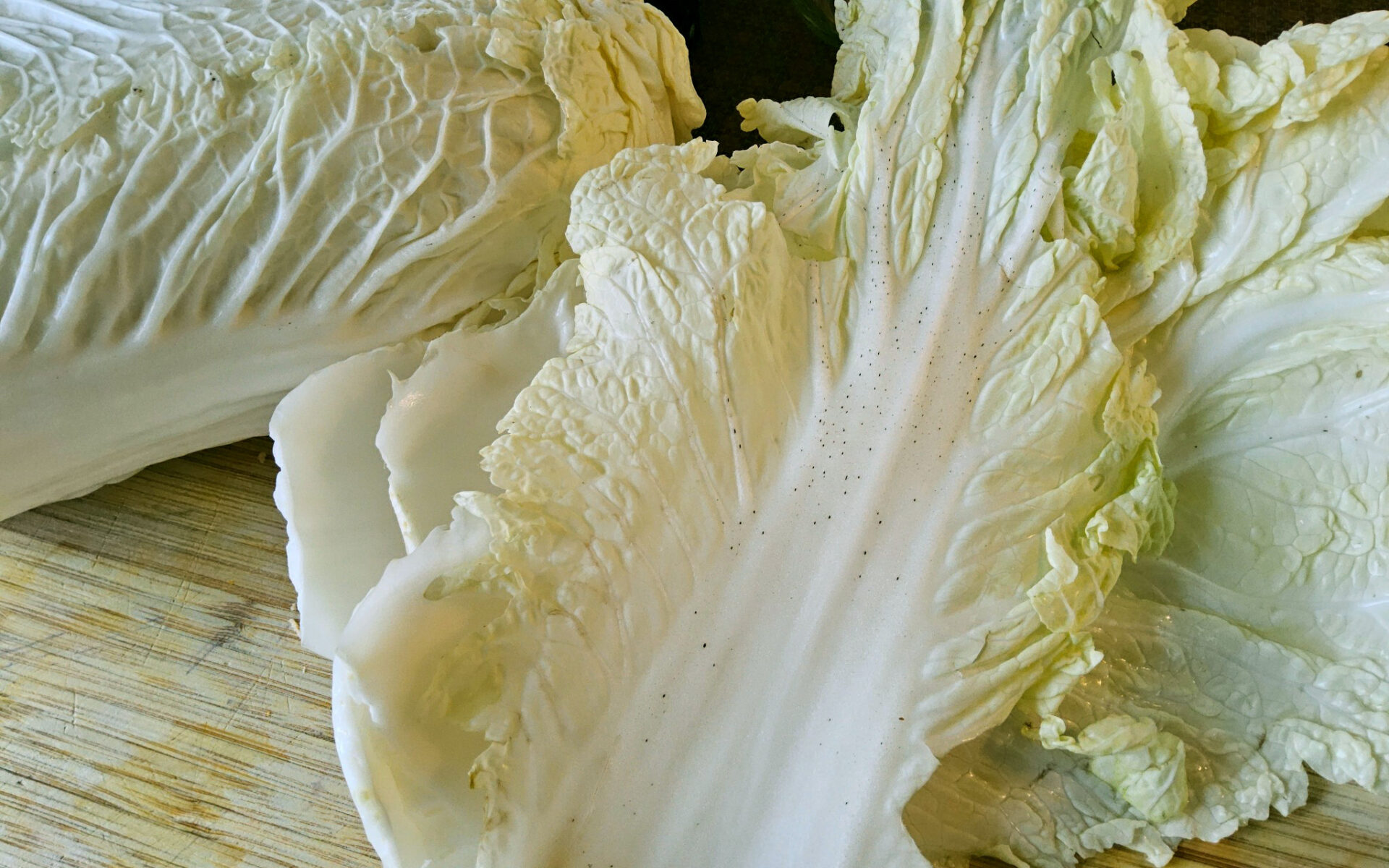 Nahaufnahme eines Chinakohlblatts, das deutliche Anzeichen von kleinen, dunklen "Pfeffenflecken" (physiologische Störung) auf der weißen Mittelrippe und den hellgrünen Bereichen aufweist. Im Hintergrund sind weitere Blätter des Chinakohls auf einem Holzschneidebrett zu sehen.