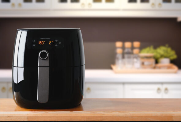 Ein schwarzer Airfryer mit beleuchtetem Bedienfeld steht auf einer Holzarbeitsplatte in einer hellen Küche.