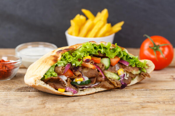 Gefüllte Dönertasche mit Salat, Tomaten, Gurken und Dönerfleisch, im Hintergrund Pommes und Soßen.