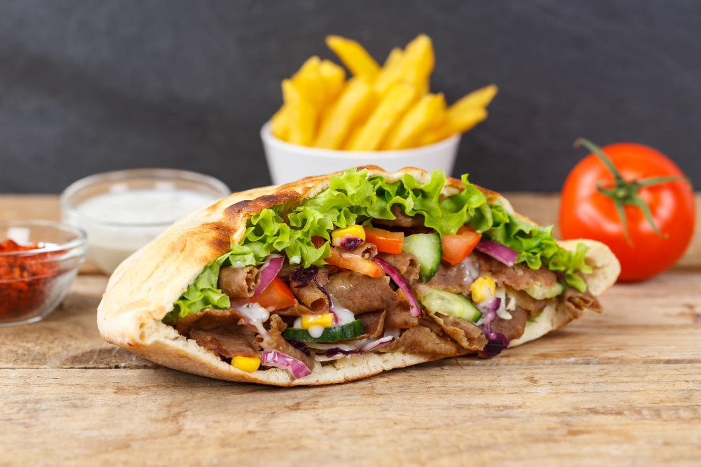 Gefüllte Dönertasche mit Salat, Tomaten, Gurken und Dönerfleisch, im Hintergrund Pommes und Soßen.