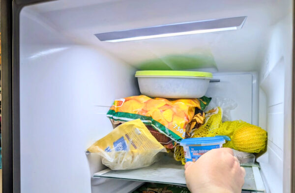 Offenes Gefrierfach mit verschiedenen Lebensmitteln, darunter eine Plastikdose mit grünem Deckel, gefrorene Pommes im Beutel, Zitrusfrüchte im gelben Netz und eine Hand, die einen Senfbecher ins Fach stellt.