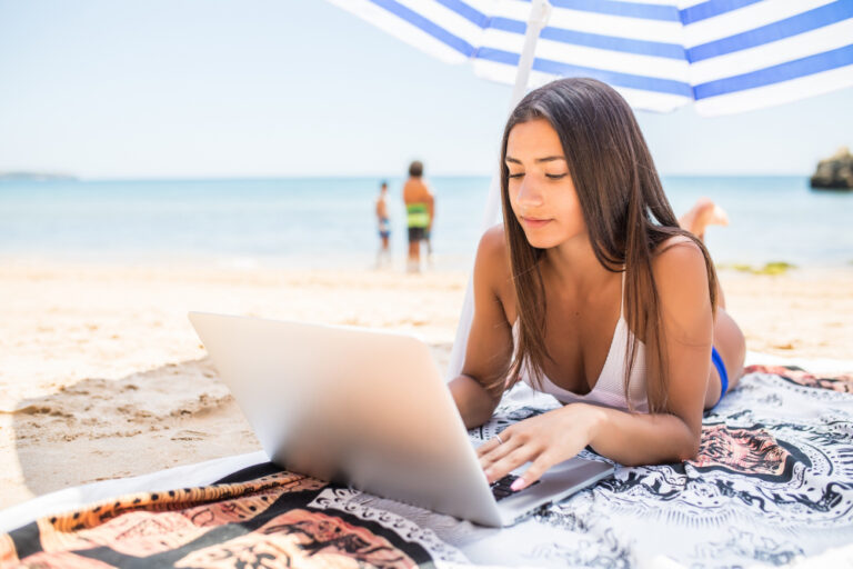Frau liegt unter Sonnenschirm am Strand und surft am Laptop.