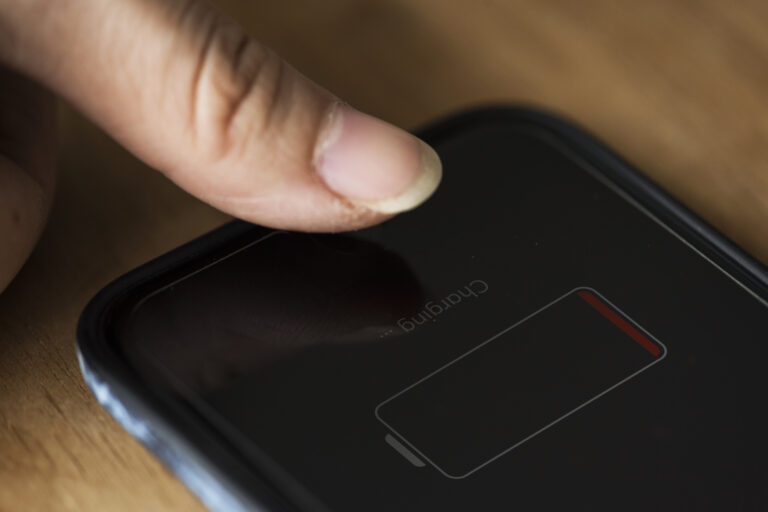 Ein Finger berührt ein schwarzes Smartphone auf einer Holzfläche; auf dem dunklen Display ist ein fast leerer Akku zu sehen.