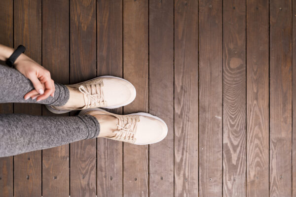 Von oben: Eine Person in grauen Leggings und hellen Sportschuhen steht auf dunklen Holzdielen; eine Hand liegt am Bein.