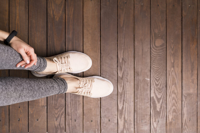 Von oben: Eine Person in grauen Leggings und hellen Sportschuhen steht auf dunklen Holzdielen; eine Hand liegt am Bein.