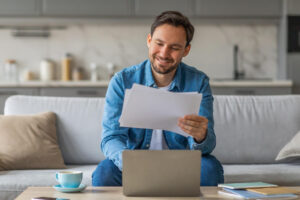 Mann sitzt auf dem Sofa mit Laptop und Steuerunterlagen in der Hand