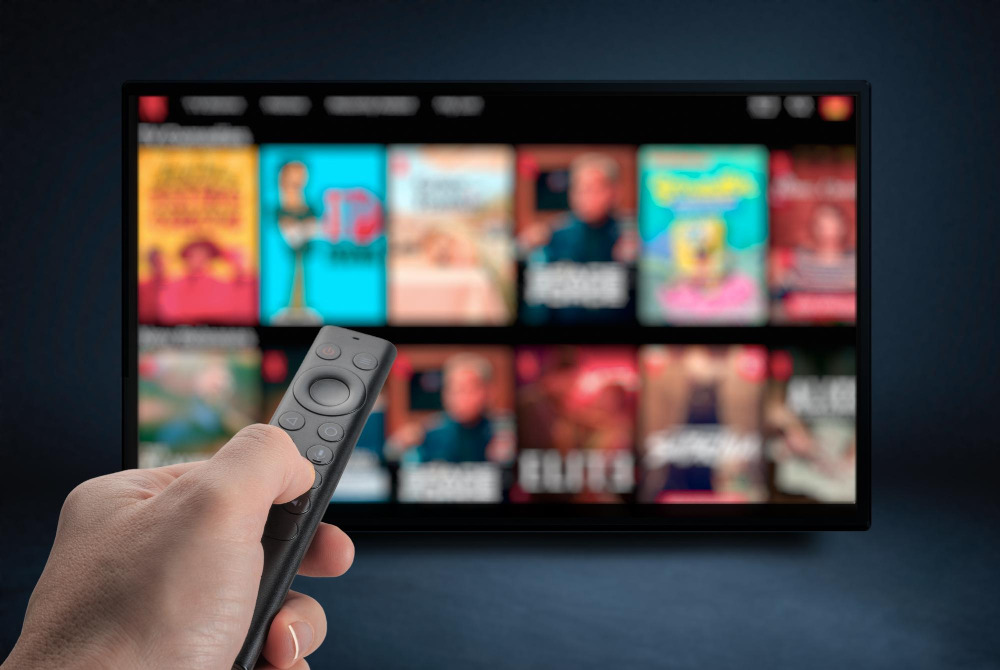 Streaming-Hopping: Hand mit Fernbedienung vor einem Fernseher mit unscharfer Streaming-Auswahl.